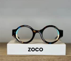 Lunettes rondes écaillées ZOCO avec cerclage intérieur bleu et jaune sur un socle blanc.