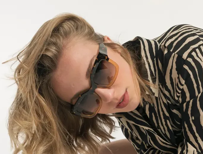 Femme blonde stylée en lunettes de soleil bicolores et chemise zébrée posant sur blanc.