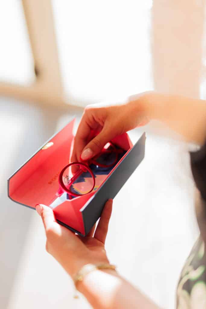 Lunettes rouges rondes Origine France Garantie en étui Mains tenant des lunettes rouges rondes 'Origine France Garantie' dans un étui chic bicolore.